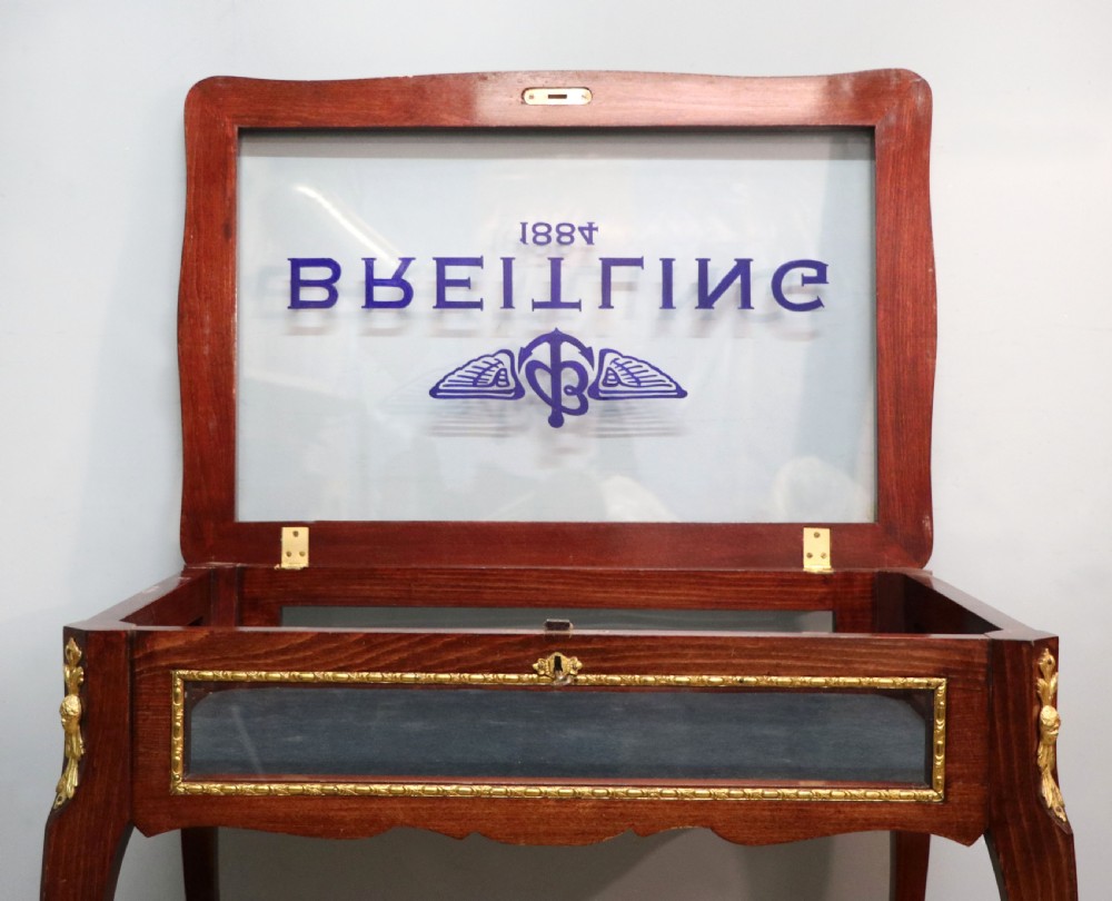 A French Louis XV style glazed bijouterie table in dark hardwood with cabriole legs, gilt metal mounts, and blue Breitling lettering on the glass top. View XVII