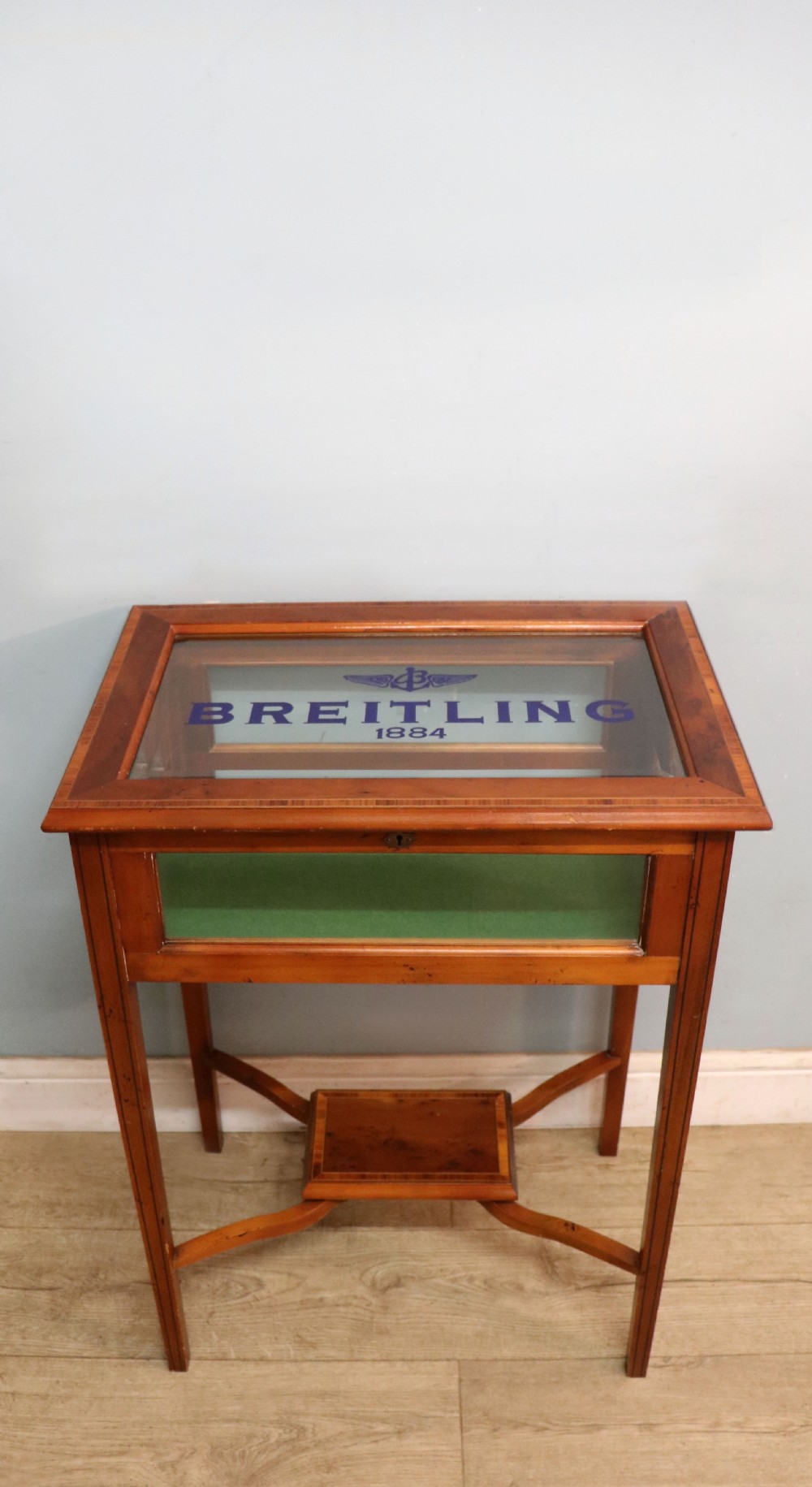 A glazed yew wood bijouterie display table featuring a green velvet interior lining, lower undertier, and Breitling 1884 logo printed on the top glass.