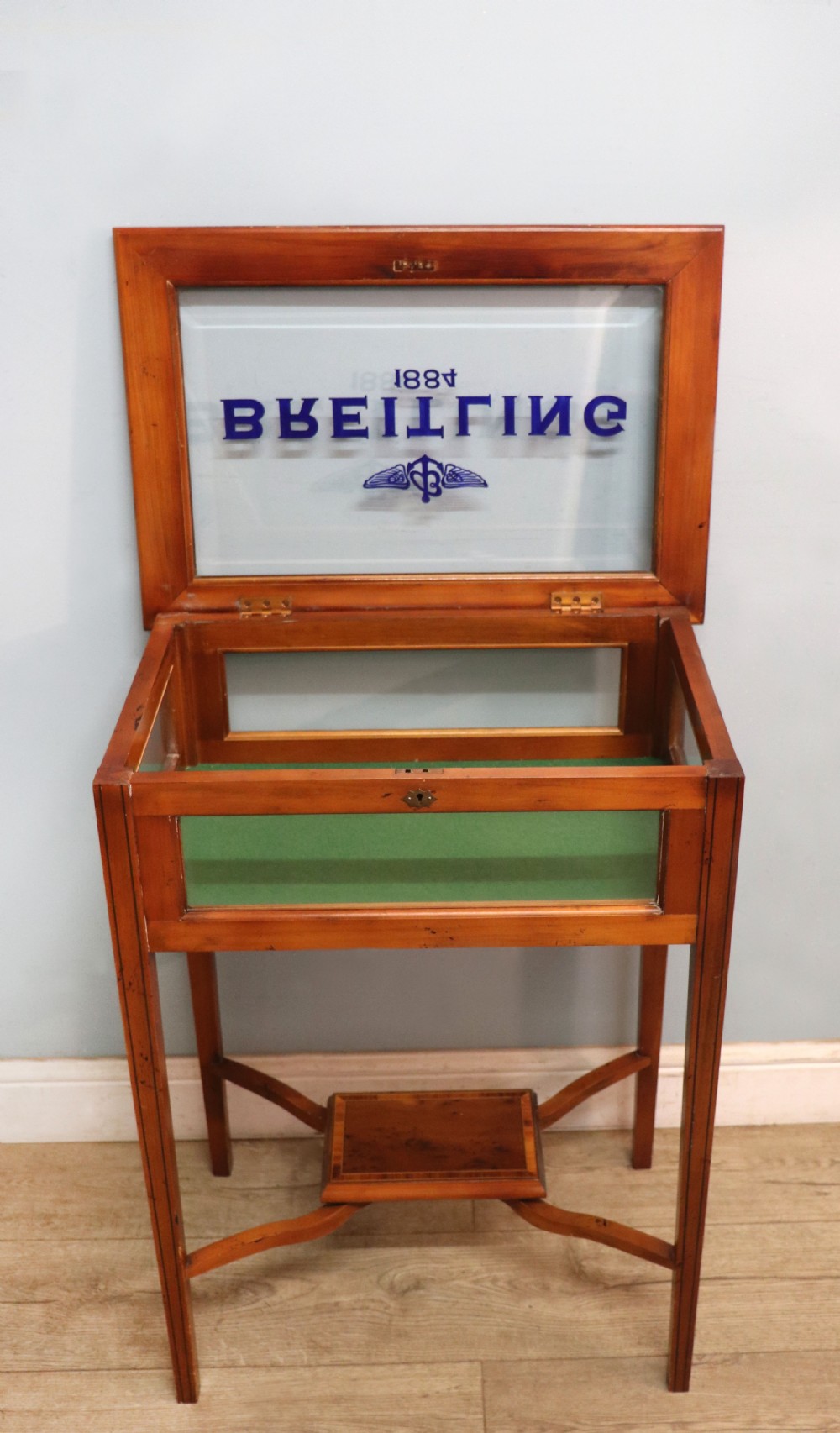 A glazed yew wood bijouterie display table featuring a green velvet interior lining, lower undertier, and Breitling 1884 logo printed on the top glass. View IV