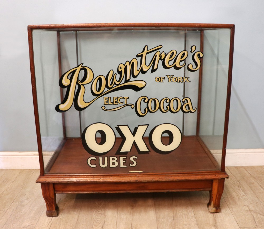 A glazed hardwood countertop display cabinet with sign-written lettering for Rowntrees Elect Cocoa and OXO Cubes on the front glass panel.