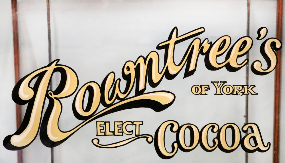A glazed hardwood countertop display cabinet with sign-written lettering for Rowntrees Elect Cocoa and OXO Cubes on the front glass panel. View X