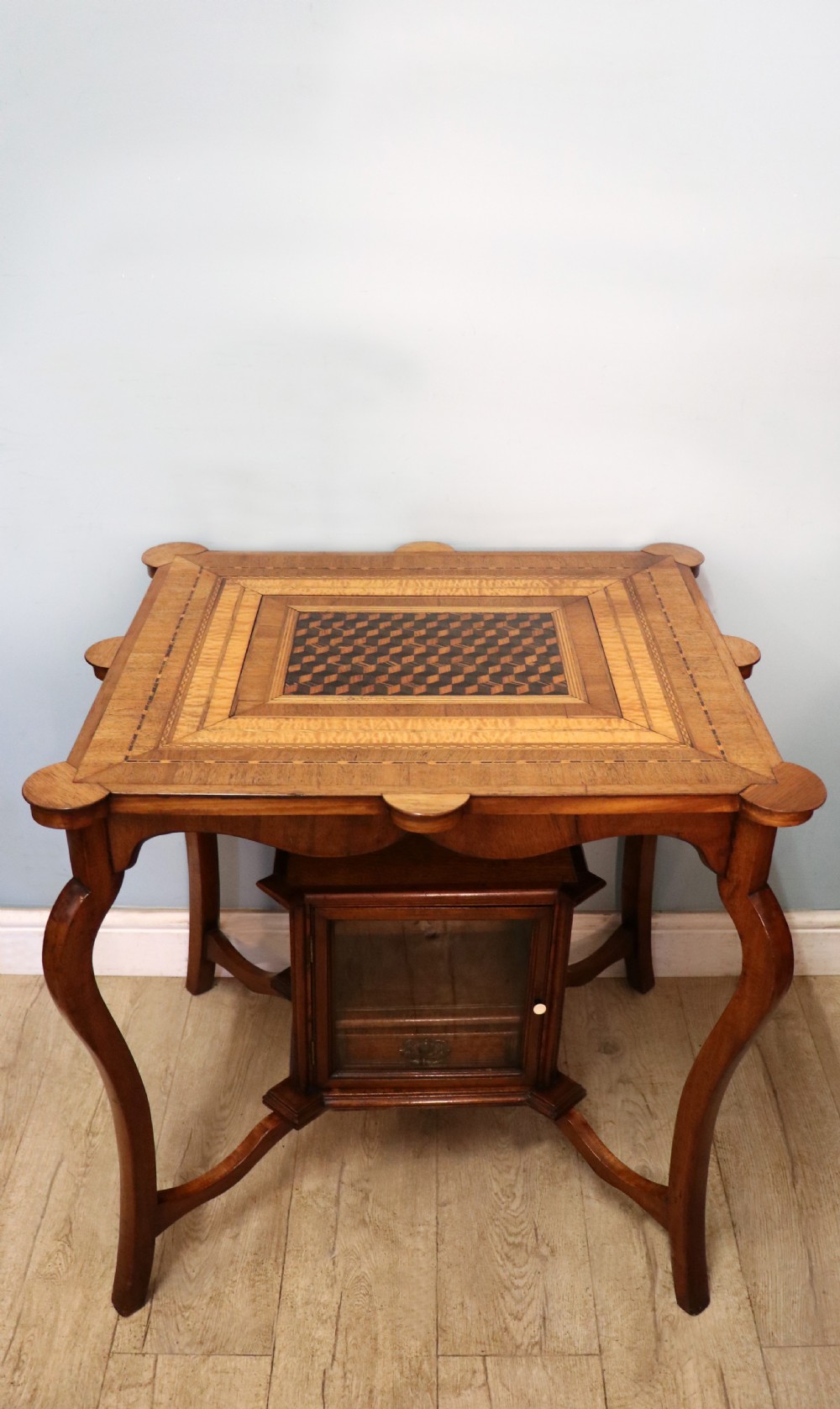 A late Victorian wooden occasional table featuring a geometric parquetry inlaid top, slender cabriole legs, and a lower glazed smokers cabinet.