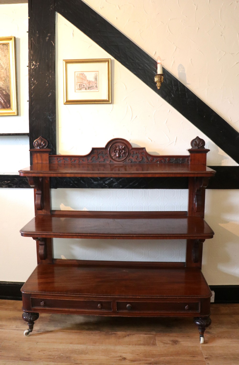A Victorian mahogany three-tiered dumb waiter featuring carved decorative pediments, supporting brackets, lower drawers, and ceramic castors. View II