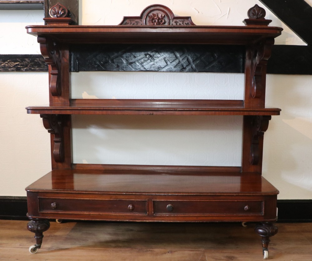 A Victorian mahogany three-tiered dumb waiter featuring carved decorative pediments, supporting brackets, lower drawers, and ceramic castors.