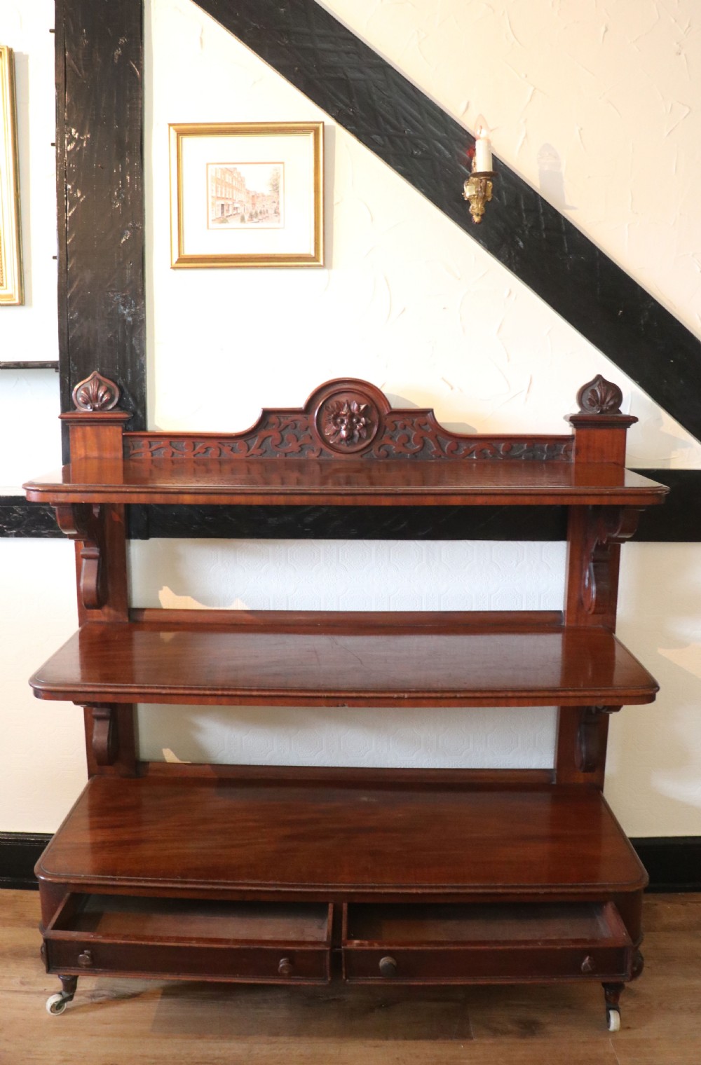 A Victorian mahogany three-tiered dumb waiter featuring carved decorative pediments, supporting brackets, lower drawers, and ceramic castors. View III