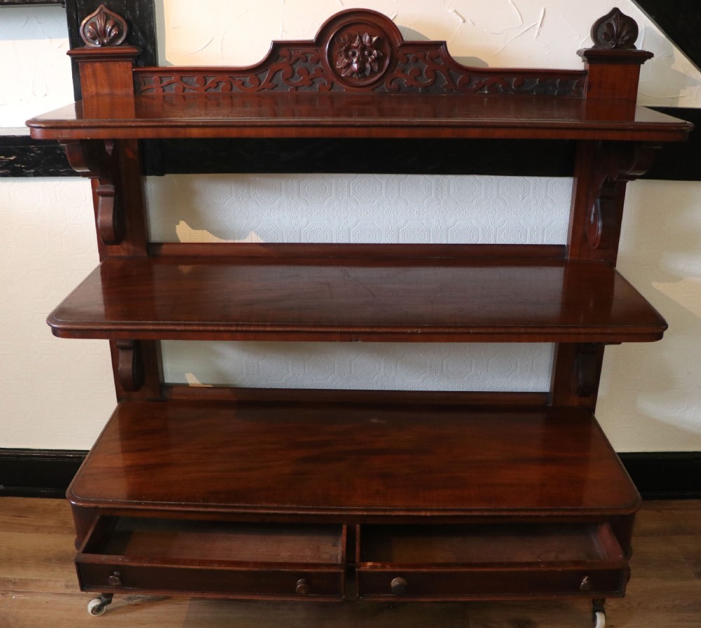 A Victorian mahogany three-tiered dumb waiter featuring carved decorative pediments, supporting brackets, lower drawers, and ceramic castors. View IV