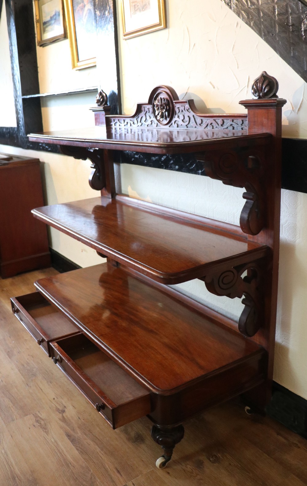 A Victorian mahogany three-tiered dumb waiter featuring carved decorative pediments, supporting brackets, lower drawers, and ceramic castors. View V