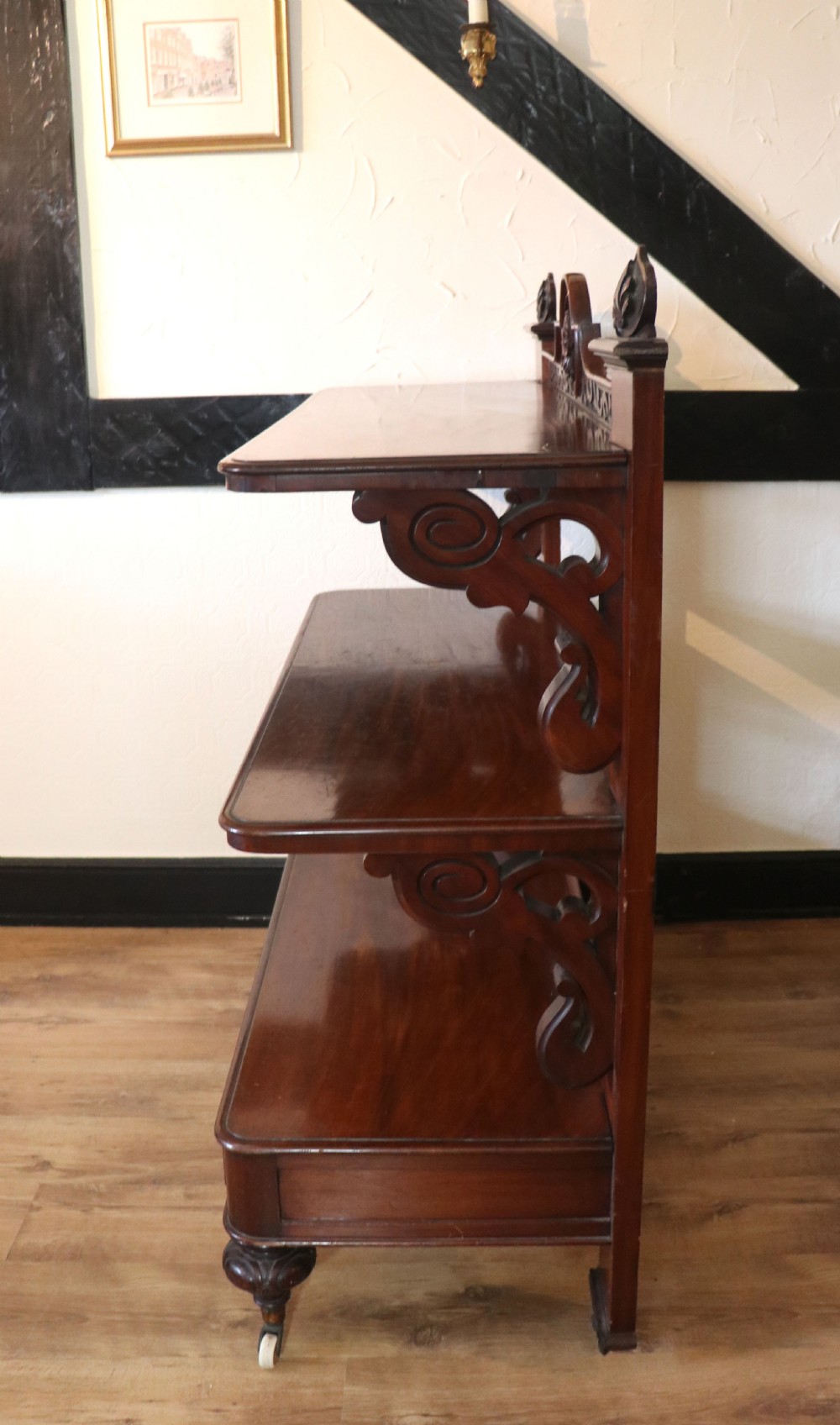 A Victorian mahogany three-tiered dumb waiter featuring carved decorative pediments, supporting brackets, lower drawers, and ceramic castors. View VI