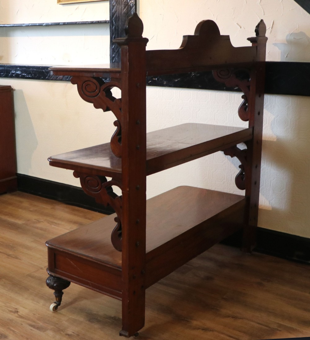 A Victorian mahogany three-tiered dumb waiter featuring carved decorative pediments, supporting brackets, lower drawers, and ceramic castors. View VII