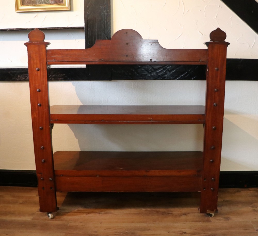 A Victorian mahogany three-tiered dumb waiter featuring carved decorative pediments, supporting brackets, lower drawers, and ceramic castors. View VIII