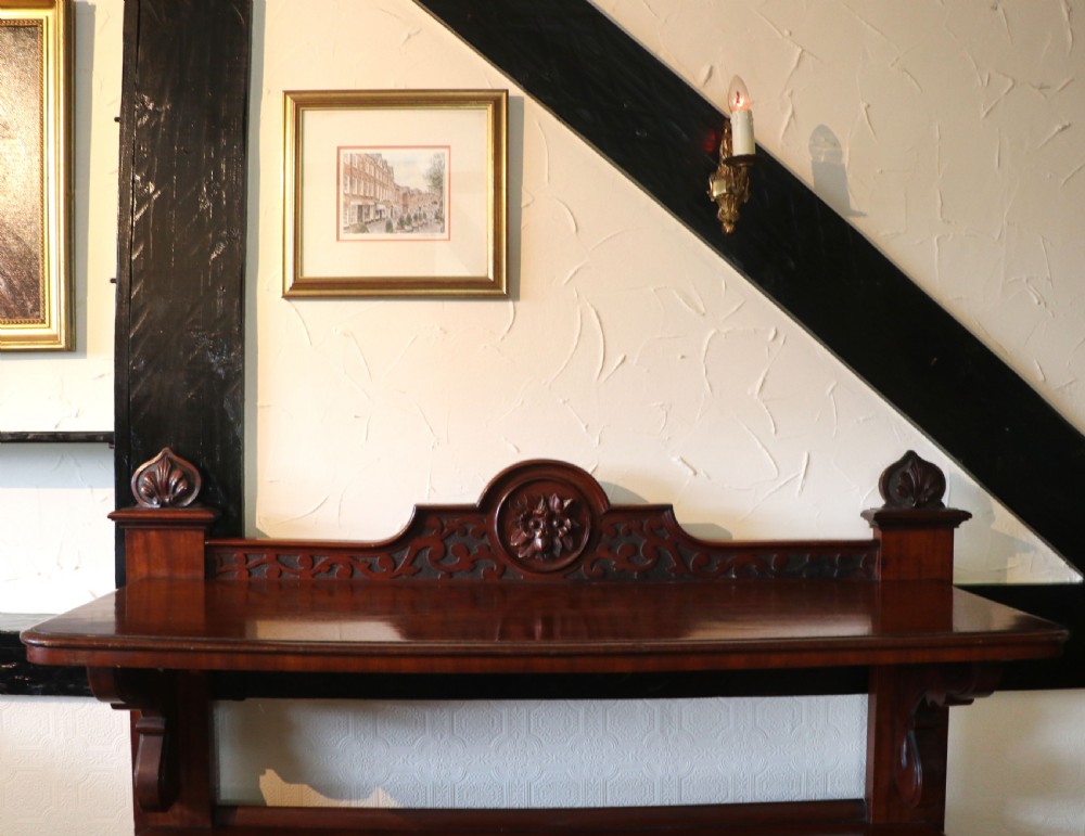 A Victorian mahogany three-tiered dumb waiter featuring carved decorative pediments, supporting brackets, lower drawers, and ceramic castors. View IX