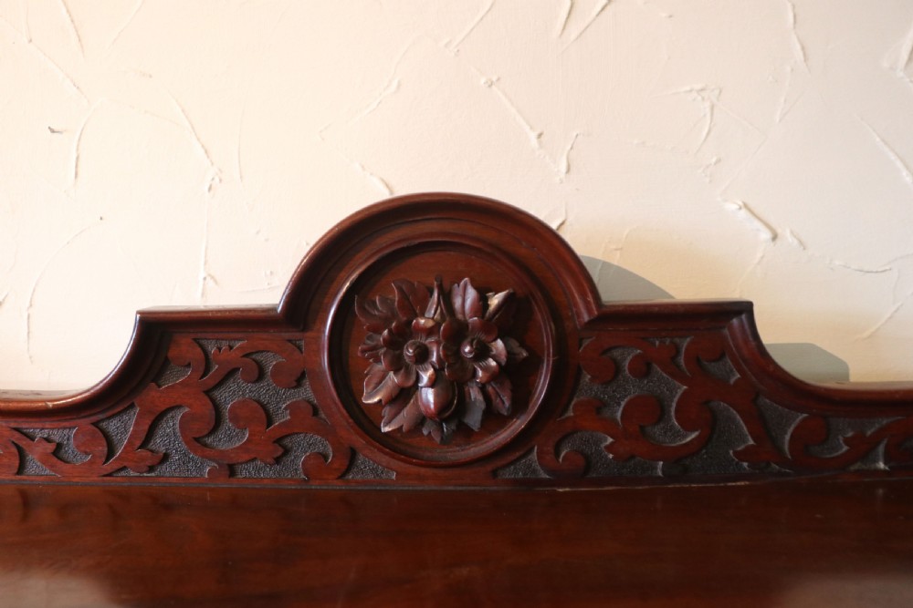 A Victorian mahogany three-tiered dumb waiter featuring carved decorative pediments, supporting brackets, lower drawers, and ceramic castors. View XII