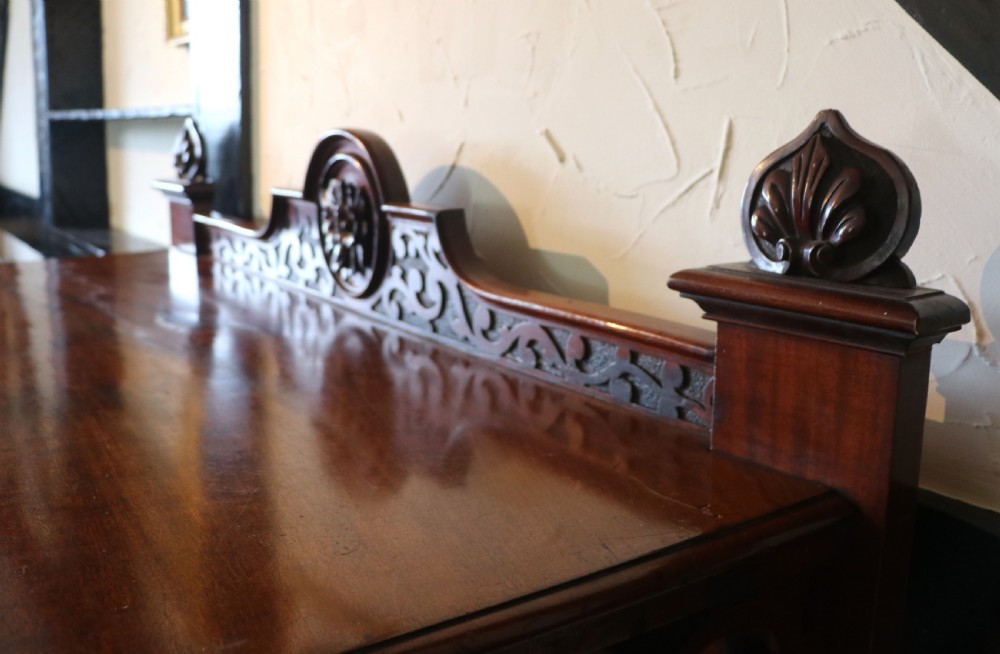 A Victorian mahogany three-tiered dumb waiter featuring carved decorative pediments, supporting brackets, lower drawers, and ceramic castors. View XV