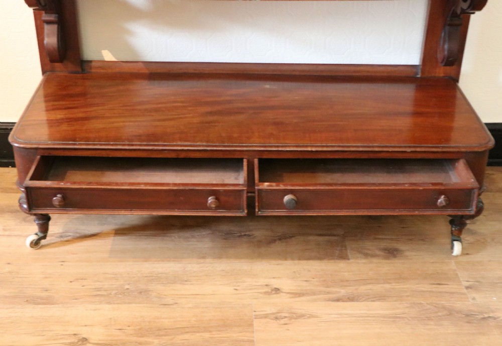 A Victorian mahogany three-tiered dumb waiter featuring carved decorative pediments, supporting brackets, lower drawers, and ceramic castors. View XX