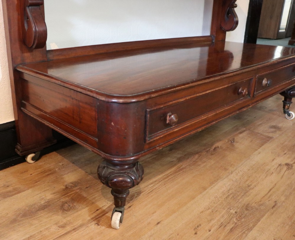 A Victorian mahogany three-tiered dumb waiter featuring carved decorative pediments, supporting brackets, lower drawers, and ceramic castors. View XXI