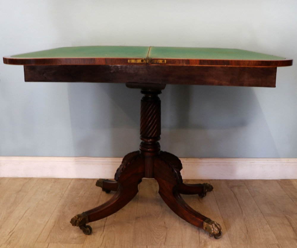 A late Georgian dark rosewood card table featuring an open fold-over green baize top supported on a spiral-carved pedestal and splayed legs with brass paw castors.