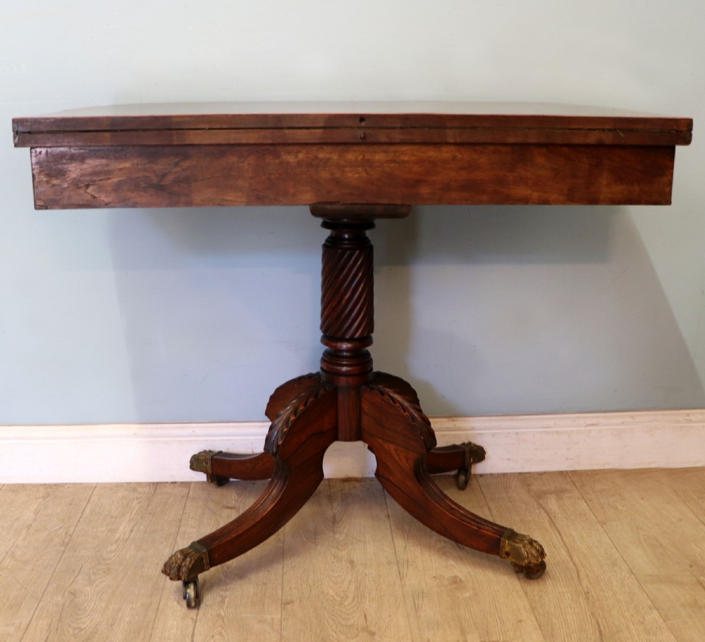 A late Georgian dark rosewood card table featuring an open fold-over green baize top supported on a spiral-carved pedestal and splayed legs with brass paw castors. View X