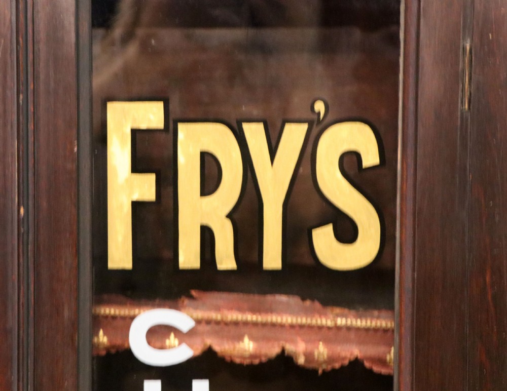 A dark mahogany Victorian display cabinet featuring glazed doors painted with Frys Cocoa and Chocolate advertising typography. View V