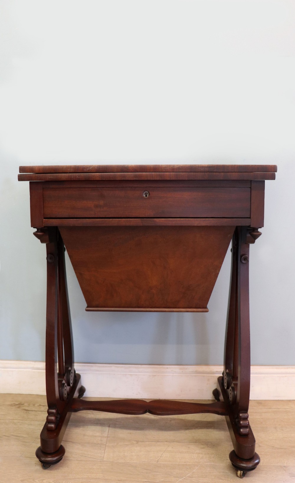 A nineteenth-century Victorian mahogany metamorphic sewing table featuring a hinged top, single drawer with brass escutcheon, tapered wooden storage bin, carved side supports, and brass castors.