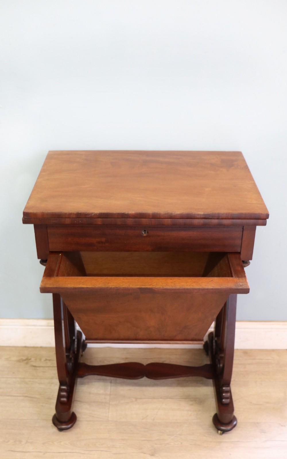 A nineteenth-century Victorian mahogany metamorphic sewing table featuring a hinged top, single drawer with brass escutcheon, tapered wooden storage bin, carved side supports, and brass castors. View IV