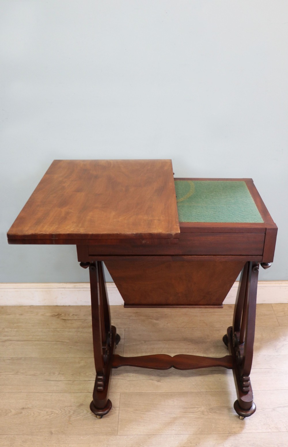 A nineteenth-century Victorian mahogany metamorphic sewing table featuring a hinged top, single drawer with brass escutcheon, tapered wooden storage bin, carved side supports, and brass castors. View VI