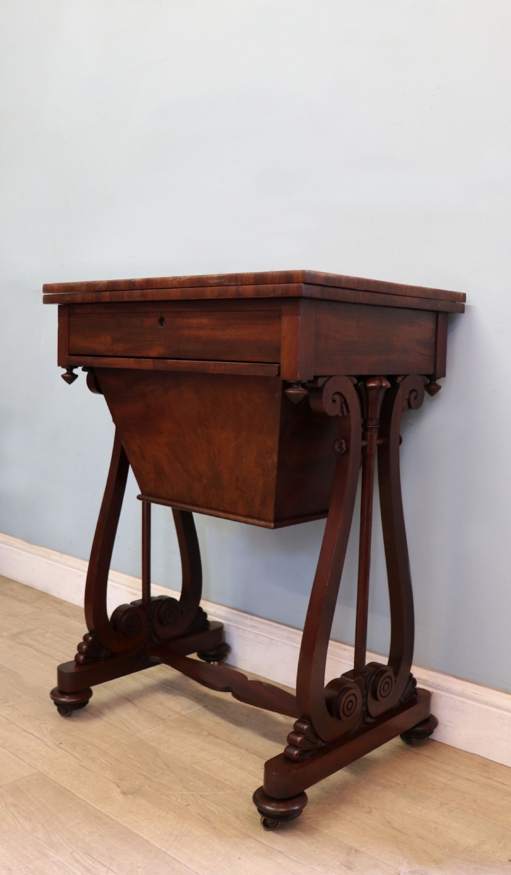 A nineteenth-century Victorian mahogany metamorphic sewing table featuring a hinged top, single drawer with brass escutcheon, tapered wooden storage bin, carved side supports, and brass castors. View IX