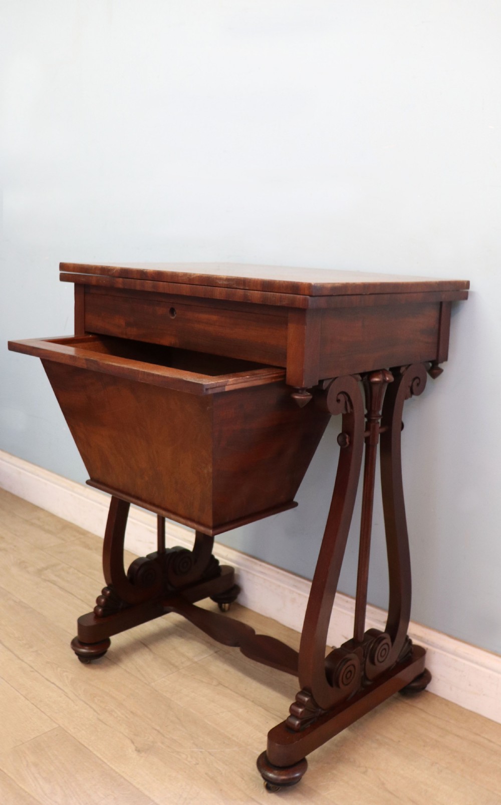 A nineteenth-century Victorian mahogany metamorphic sewing table featuring a hinged top, single drawer with brass escutcheon, tapered wooden storage bin, carved side supports, and brass castors. View X