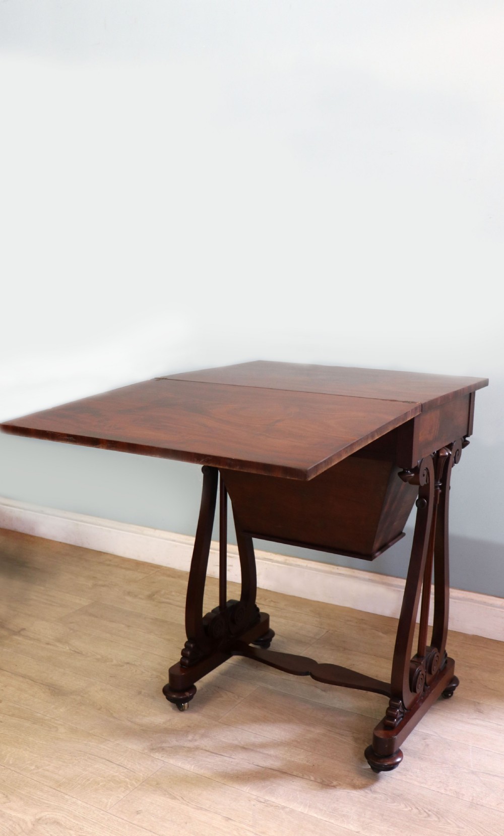 A nineteenth-century Victorian mahogany metamorphic sewing table featuring a hinged top, single drawer with brass escutcheon, tapered wooden storage bin, carved side supports, and brass castors. View XII
