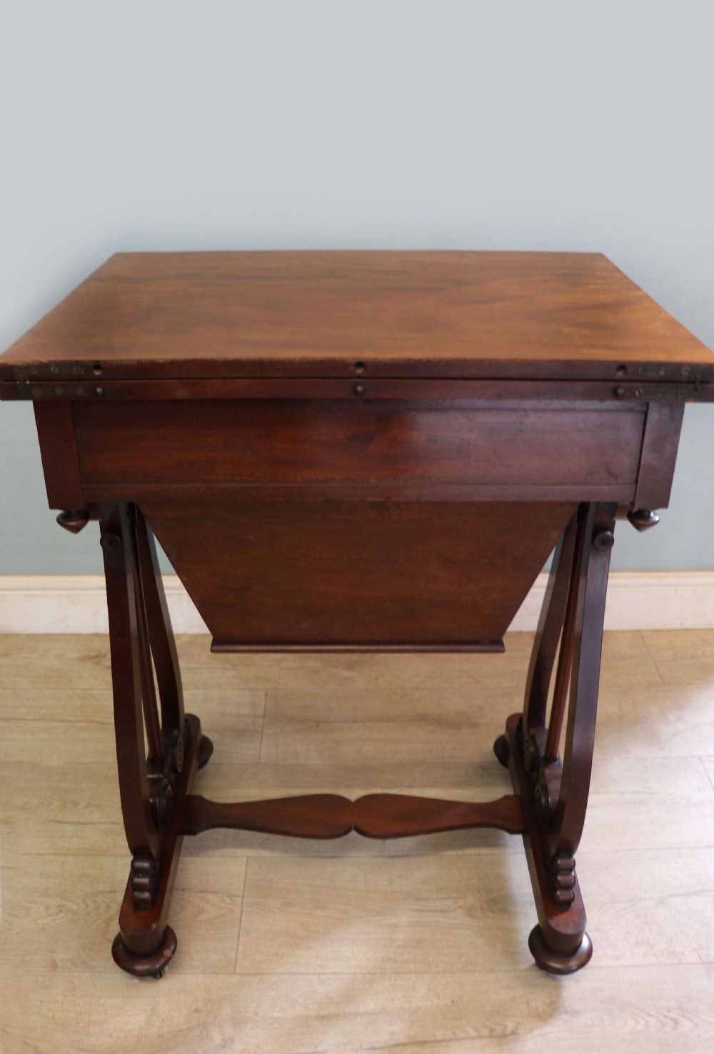 A nineteenth-century Victorian mahogany metamorphic sewing table featuring a hinged top, single drawer with brass escutcheon, tapered wooden storage bin, carved side supports, and brass castors. View XIII