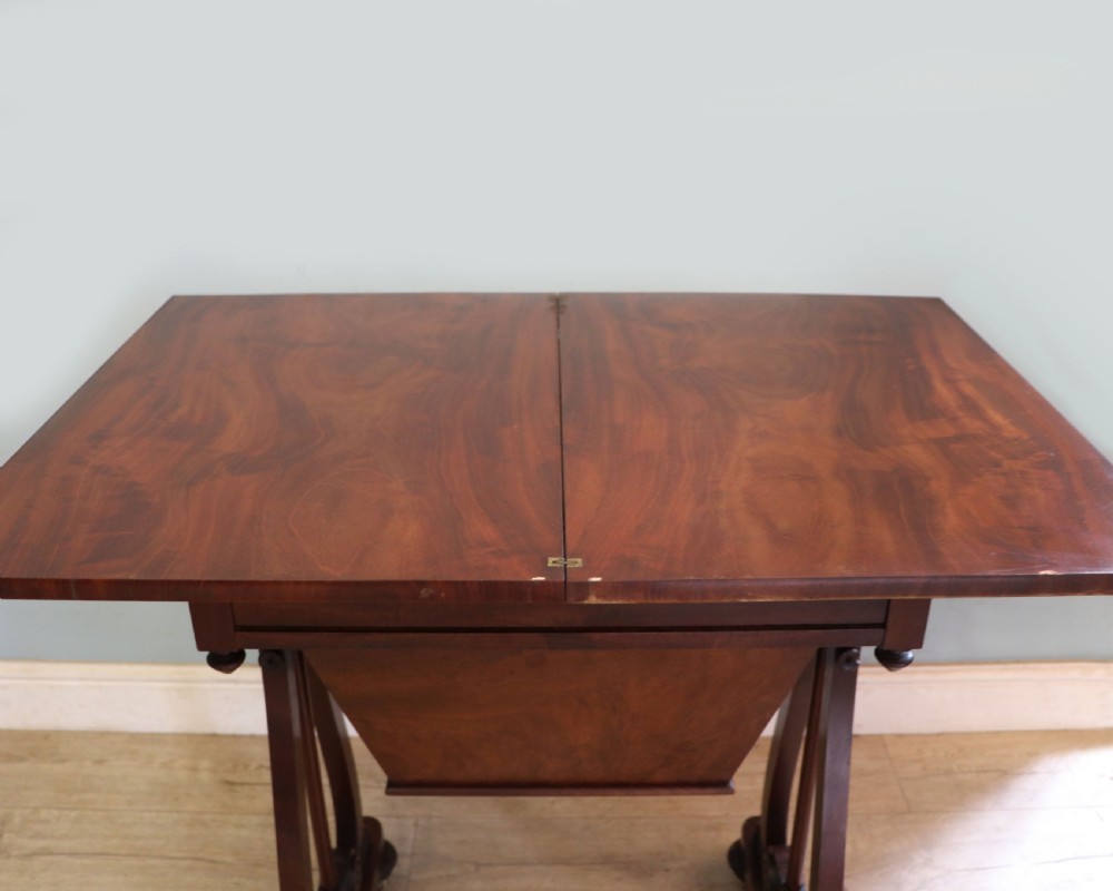 A nineteenth-century Victorian mahogany metamorphic sewing table featuring a hinged top, single drawer with brass escutcheon, tapered wooden storage bin, carved side supports, and brass castors. View XVII