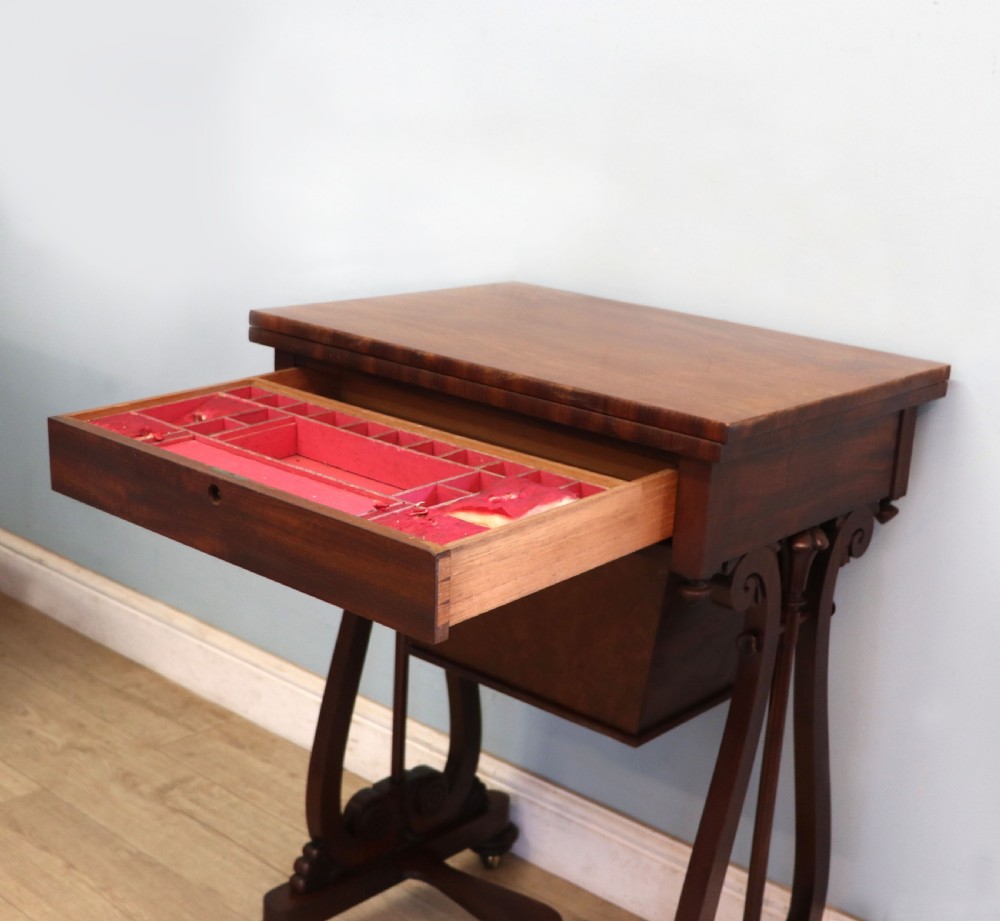 A nineteenth-century Victorian mahogany metamorphic sewing table featuring a hinged top, single drawer with brass escutcheon, tapered wooden storage bin, carved side supports, and brass castors. View XX