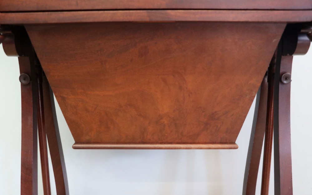 A nineteenth-century Victorian mahogany metamorphic sewing table featuring a hinged top, single drawer with brass escutcheon, tapered wooden storage bin, carved side supports, and brass castors. View XXV