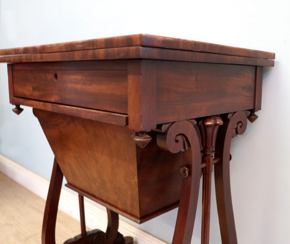 A nineteenth-century Victorian mahogany metamorphic sewing table featuring a hinged top, single drawer with brass escutcheon, tapered wooden storage bin, carved side supports, and brass castors. View XXVI