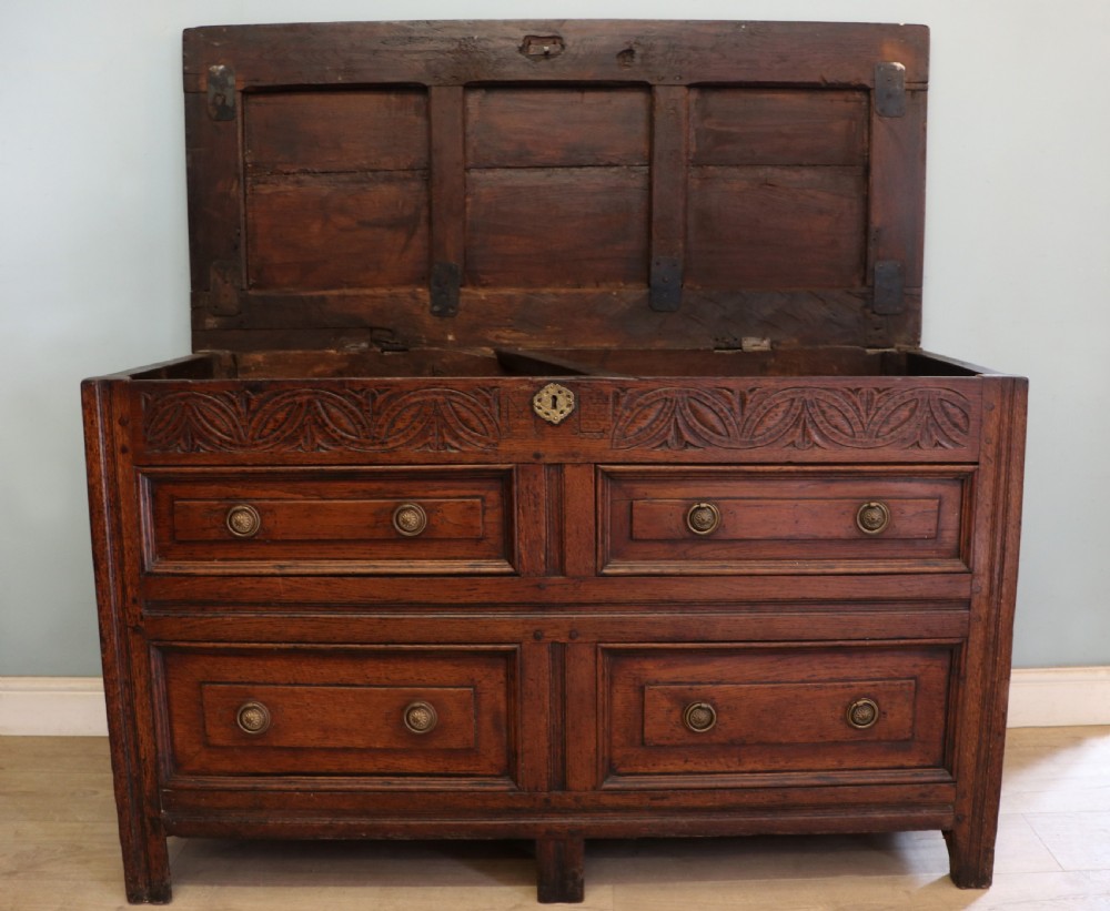 An 18th-century dark oak coffer with a hinged top lid, a carved upper frieze, and panelled lower drawers featuring brass drop handles.