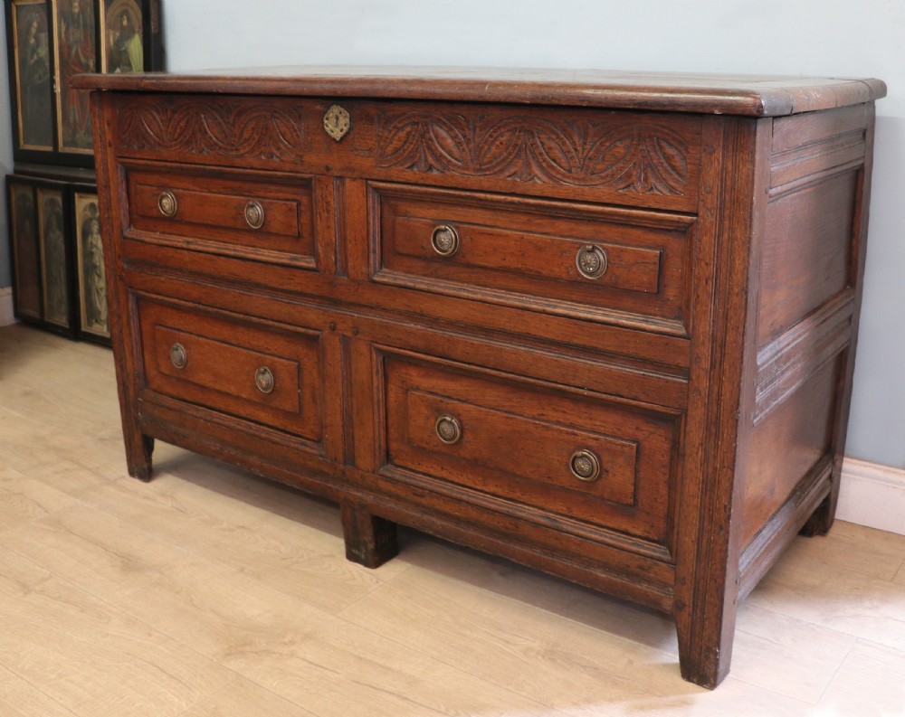 An 18th-century dark oak coffer with a hinged top lid, a carved upper frieze, and panelled lower drawers featuring brass drop handles. View IV