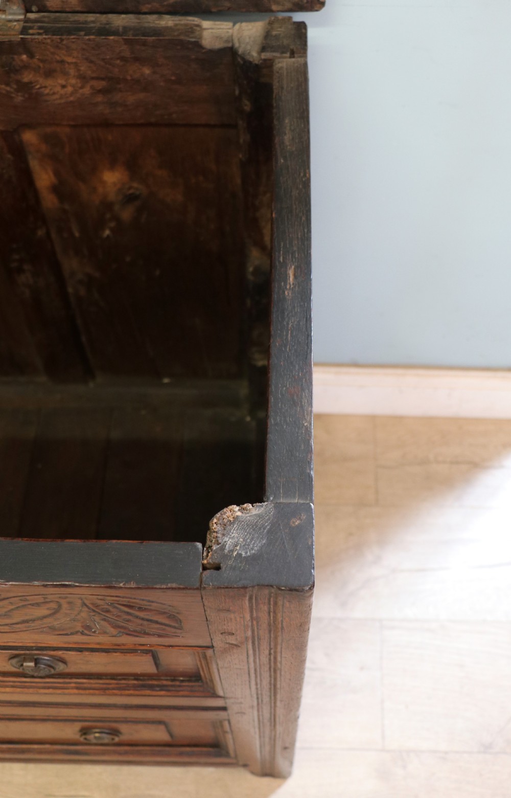 An 18th-century dark oak coffer with a hinged top lid, a carved upper frieze, and panelled lower drawers featuring brass drop handles. View XVI