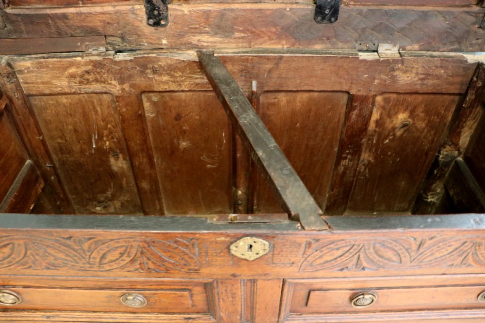 An 18th-century dark oak coffer with a hinged top lid, a carved upper frieze, and panelled lower drawers featuring brass drop handles. View XVIII