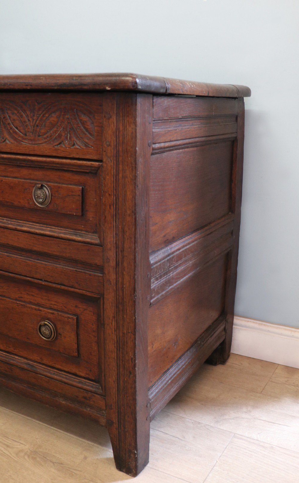 An 18th-century dark oak coffer with a hinged top lid, a carved upper frieze, and panelled lower drawers featuring brass drop handles. View XXV