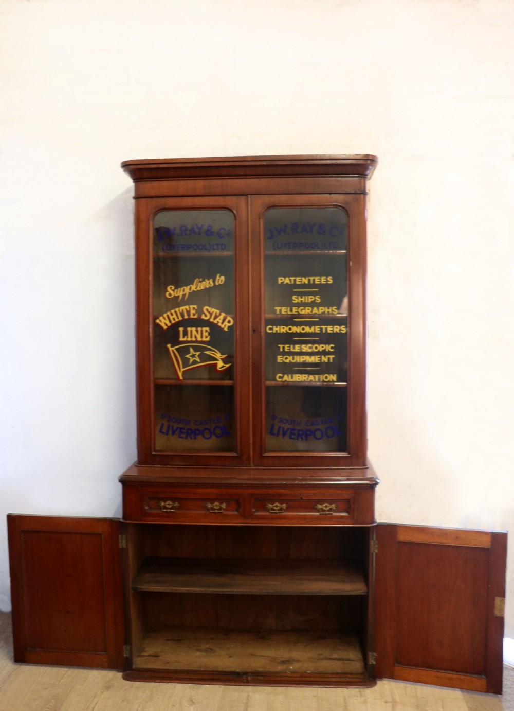 A late Victorian mahogany two-piece bookcase cabinet featuring upper glazed doors with gilded advertising lettering above two frieze drawers and a cupboard base. View V