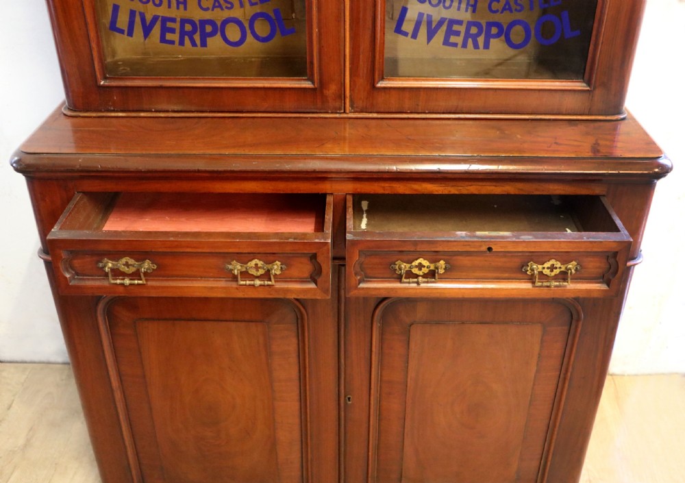 A late Victorian mahogany two-piece bookcase cabinet featuring upper glazed doors with gilded advertising lettering above two frieze drawers and a cupboard base. View XVII
