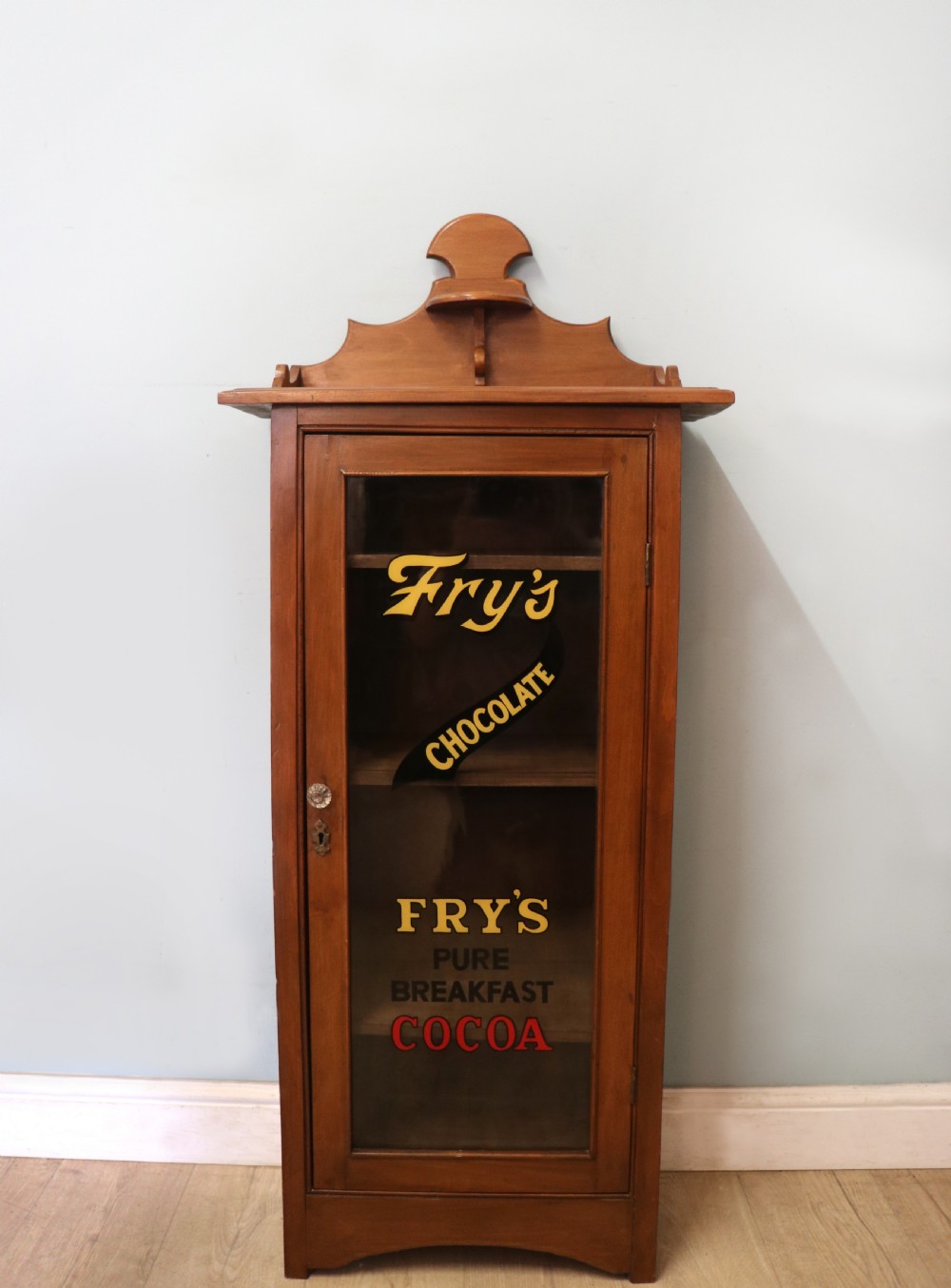 A Victorian American walnut glazed display cabinet featuring a shaped top pediment and hand-painted Frys Chocolate advertising on the single glass door.