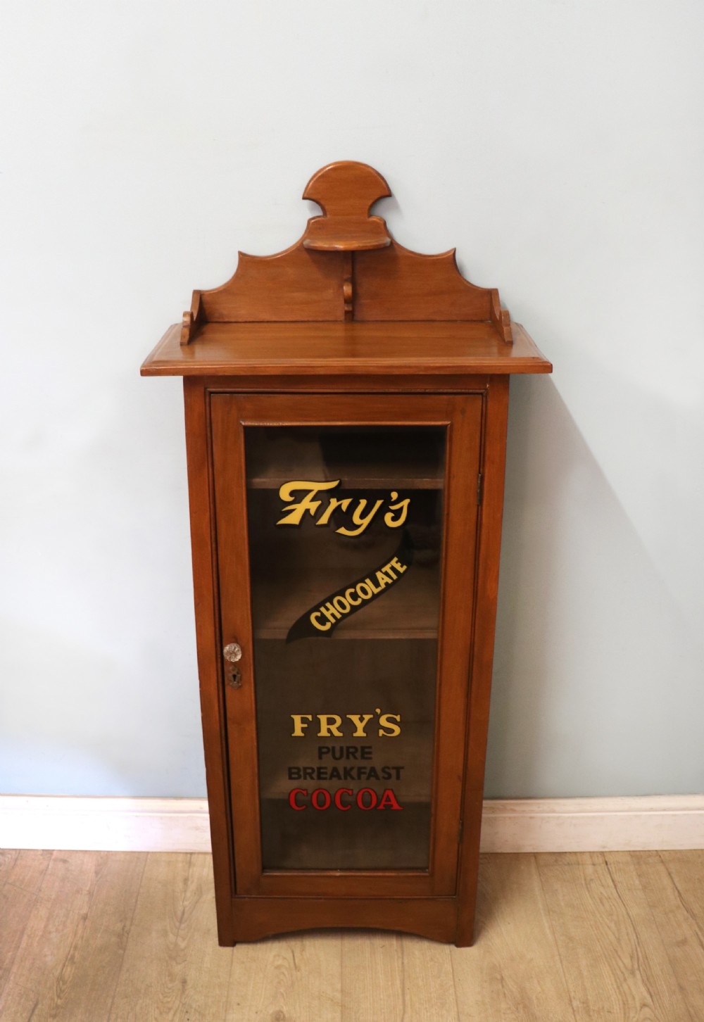 A Victorian American walnut glazed display cabinet featuring a shaped top pediment and hand-painted Frys Chocolate advertising on the single glass door. View II