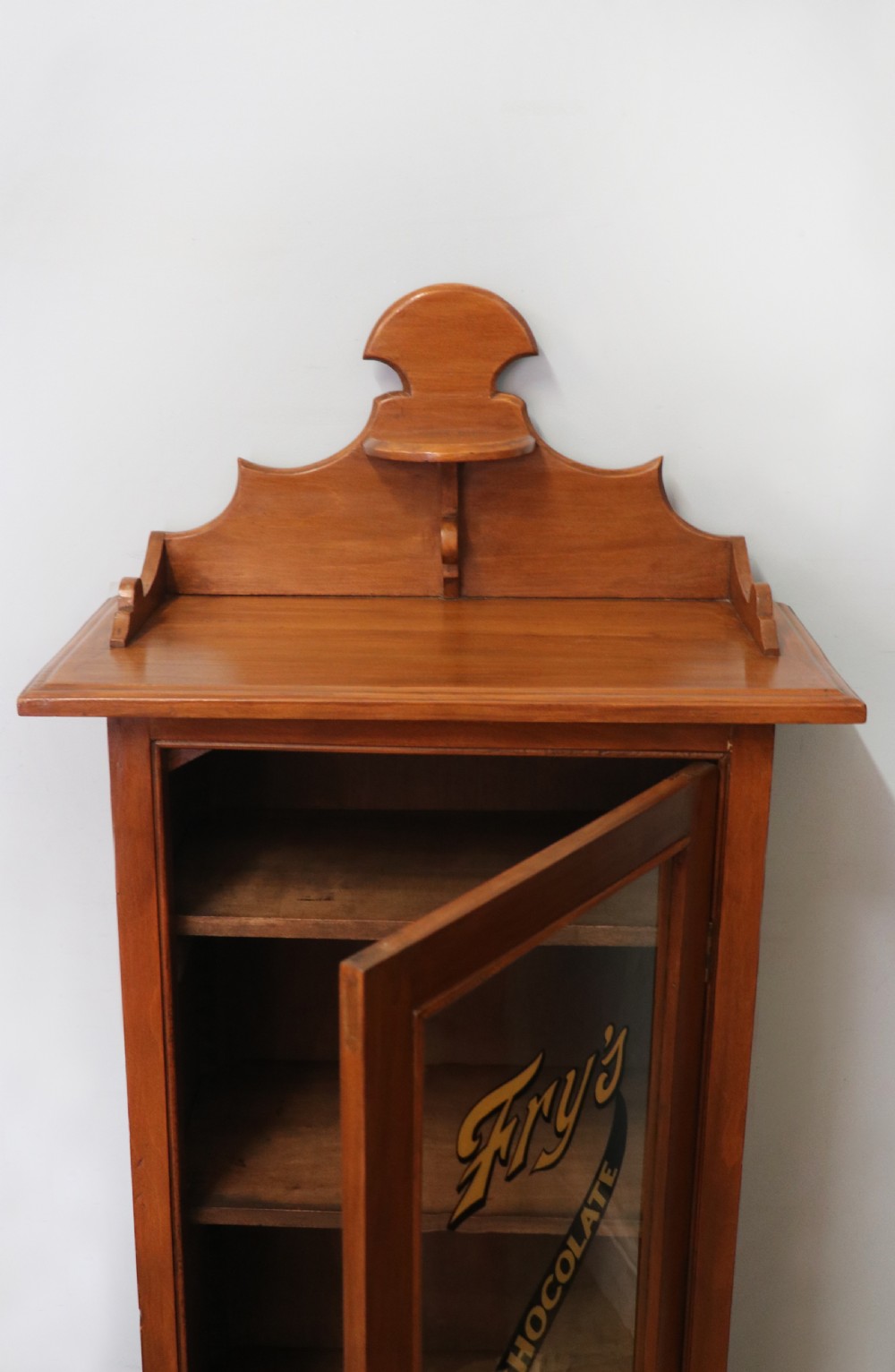 A Victorian American walnut glazed display cabinet featuring a shaped top pediment and hand-painted Frys Chocolate advertising on the single glass door. View XVIII