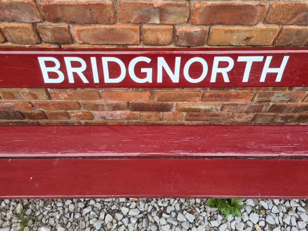 A late nineteenth to early twentieth-century painted maroon wooden railway bench featuring black cast-iron supports and white text reading BRIDGNORTH across the backrest. View V