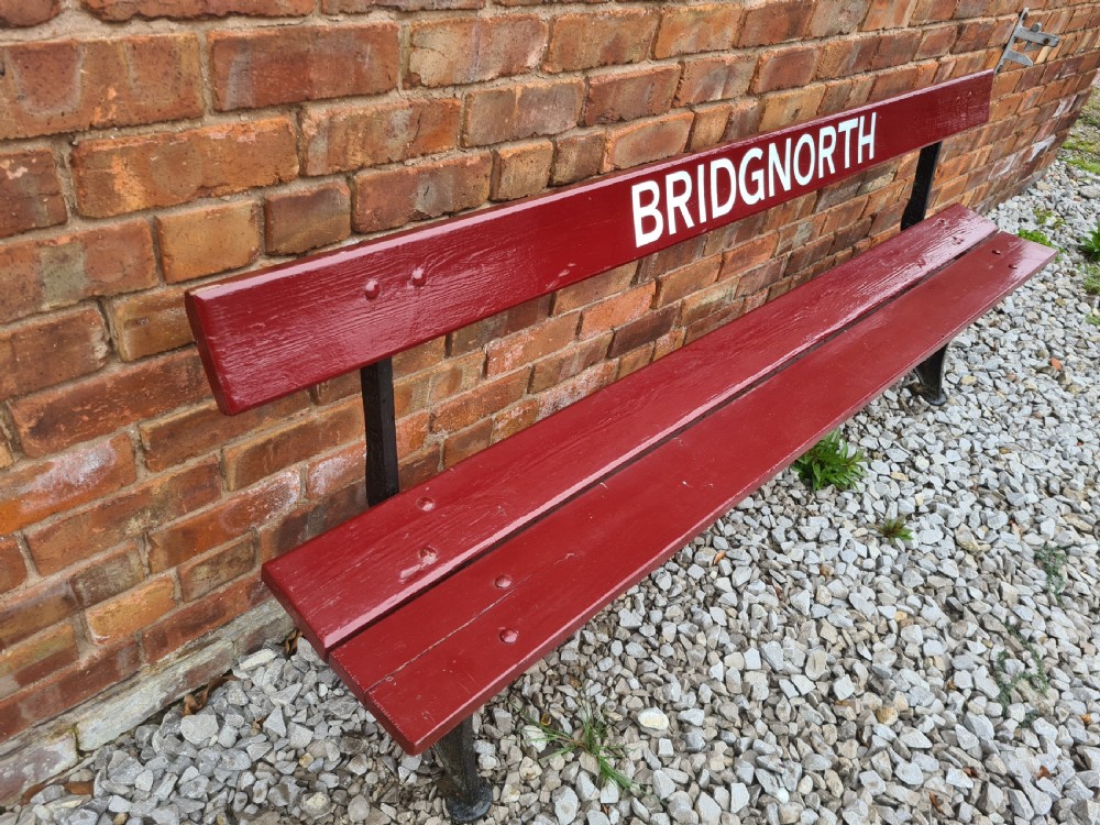 A late nineteenth to early twentieth-century painted maroon wooden railway bench featuring black cast-iron supports and white text reading BRIDGNORTH across the backrest. View VI