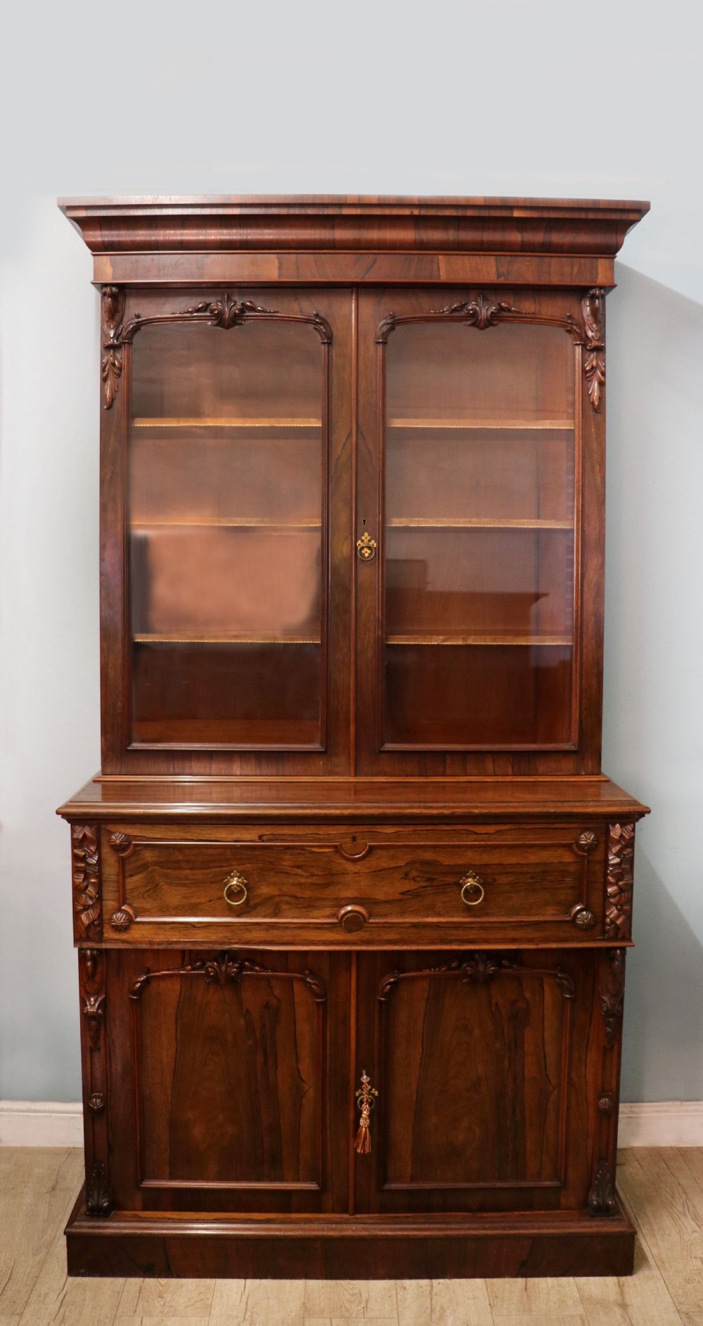 An early Victorian glazed secretaire bookcase cabinet in dark figured wood with carved arch details, a central desk drawer, and lower cupboard doors.