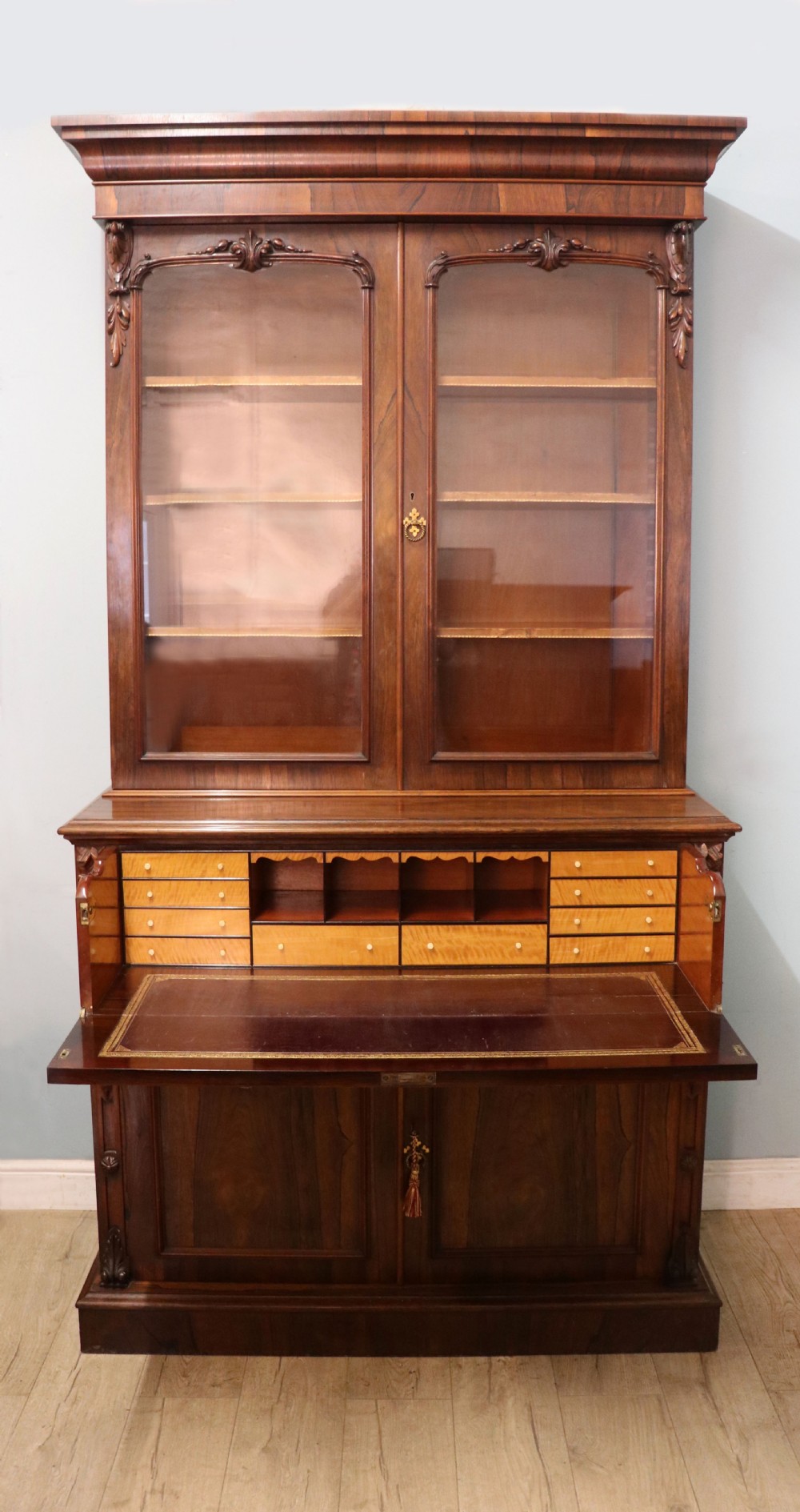 An early Victorian glazed secretaire bookcase cabinet in dark figured wood with carved arch details, a central desk drawer, and lower cupboard doors. View III