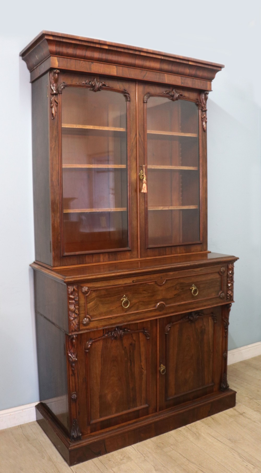 An early Victorian glazed secretaire bookcase cabinet in dark figured wood with carved arch details, a central desk drawer, and lower cupboard doors. View V