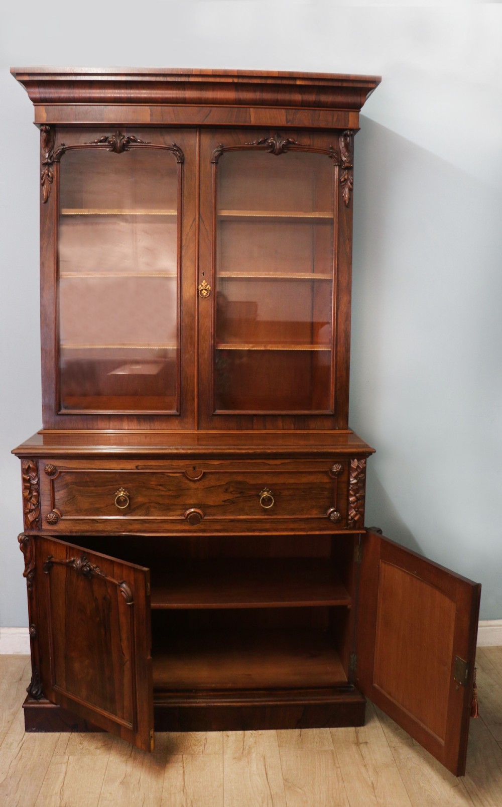 An early Victorian glazed secretaire bookcase cabinet in dark figured wood with carved arch details, a central desk drawer, and lower cupboard doors. View VI