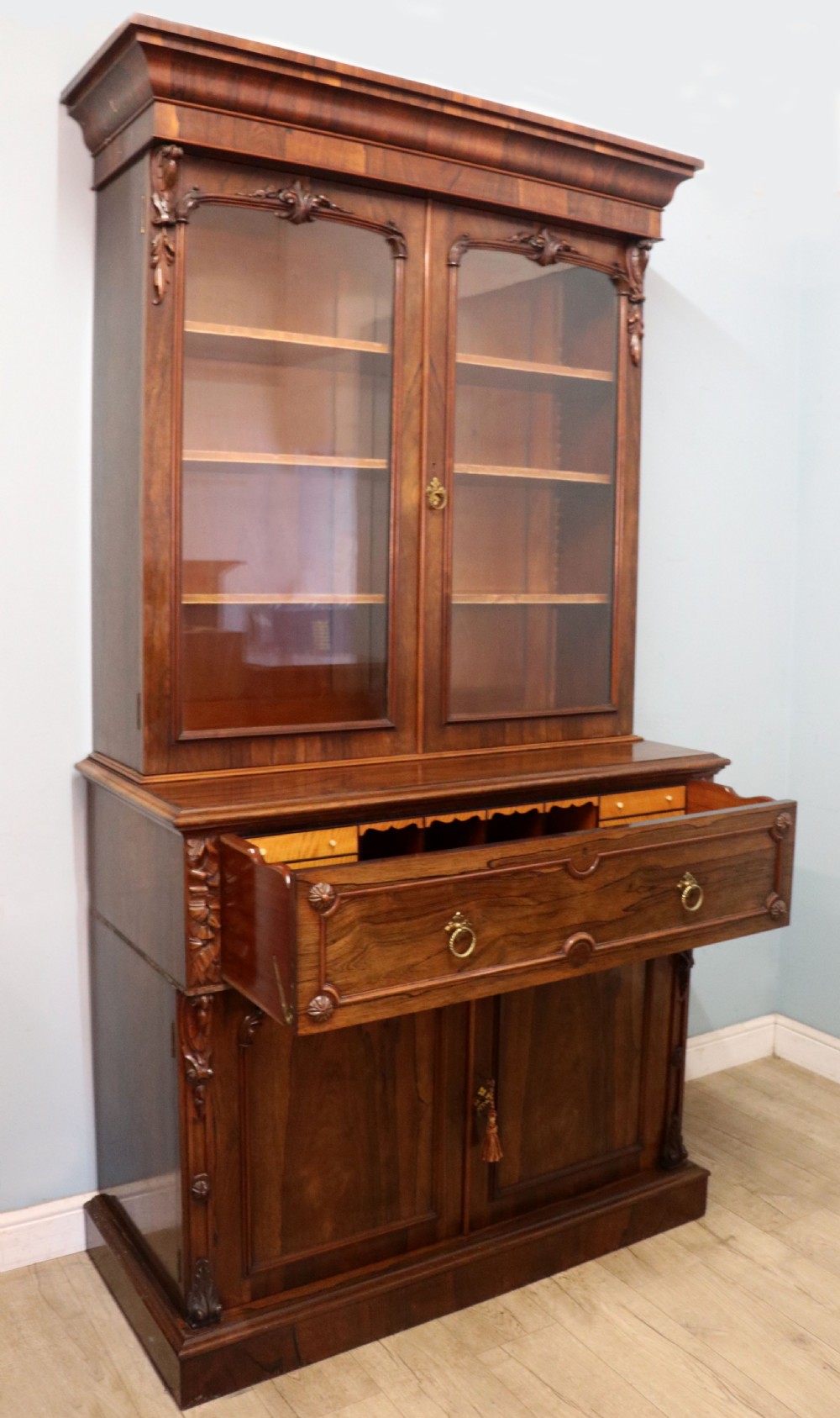 An early Victorian glazed secretaire bookcase cabinet in dark figured wood with carved arch details, a central desk drawer, and lower cupboard doors. View VII